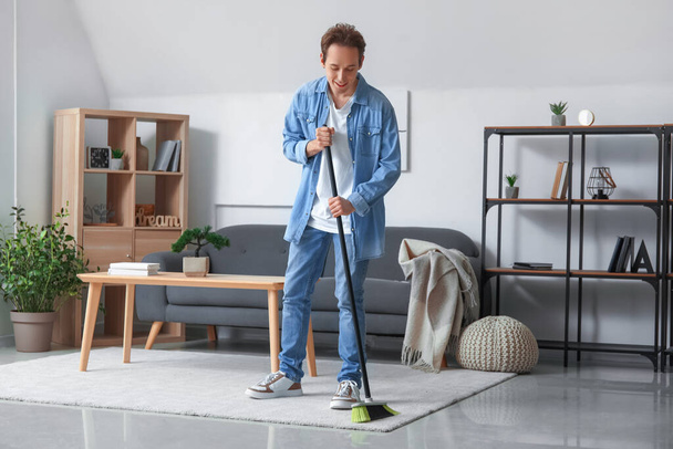 Young man sweeping carpet with broom at home - Foto, imagen