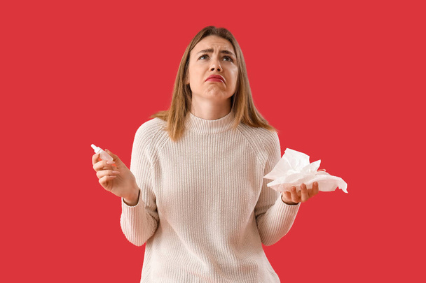 Ill young woman with nasal drops and tissues on red background - Foto, Imagen