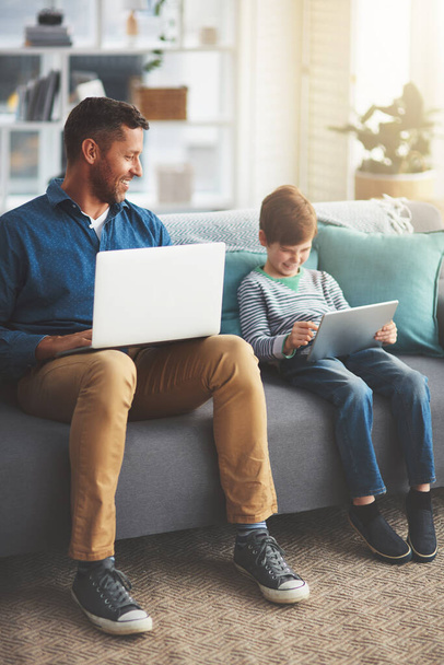 Ze werken allebei vanuit huis. een vrolijk jongetje en zijn vader met behulp van een laptop en een digitale tablet terwijl ze overdag thuis op een bank zitten - Foto, afbeelding