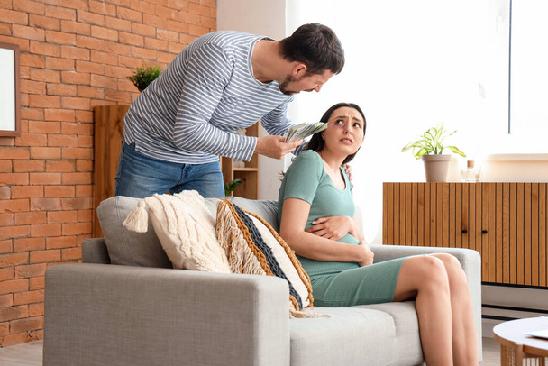 Angry husband with money and his pregnant wife at home. Domestic violence concept - Photo, Image