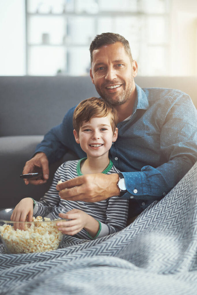 Wat een koelbloedige dag met mijn zoon. Portret van een zorgeloze jongen en zijn vader die samen een film kijken terwijl ze op de vloer zitten en overdag popcorn eten - Foto, afbeelding
