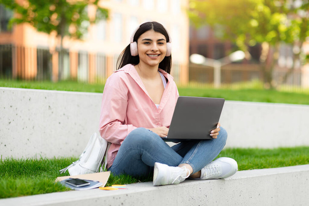 Gelukkige Spaanse student dame in draadloze koptelefoon studeren met laptop buiten, zitten met de computer op de campus, kijken en lachen naar de camera, genieten van afstandsonderwijs, kopiëren ruimte - Foto, afbeelding
