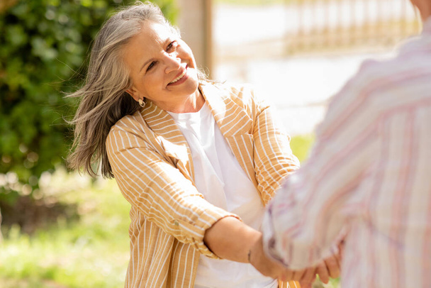 Cheerful european mature gray haired woman hold man hand, enjoy date together in park, have fun at summer weekend, outdoor. Relationship, love, romance, leisure time and walk - 写真・画像