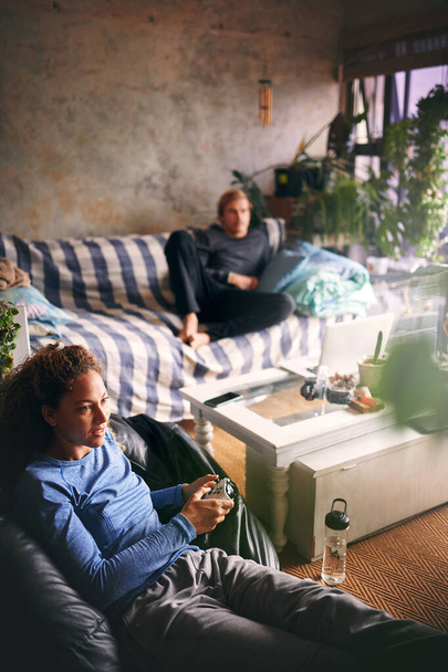 In lockdown we get our game on. a young woman playing video games while her boyfriend relaxes on the sofa in the background - Foto, Imagem