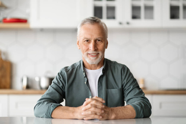 Portrait of handsome senior man sitting at table in kitchen, happy elderly gentleman in casual clothes posing in home interior, mature grey-haired male looking and smiling at camera, copy space - Photo, image
