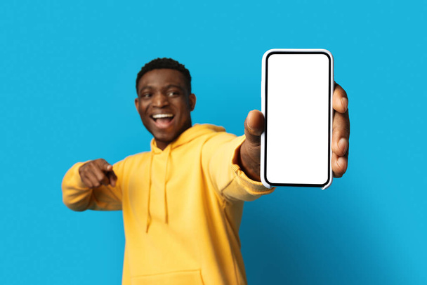 Oferta en línea. Gran teléfono celular moderno con pantalla blanca vacía en la mano del hombre afroamericano guapo feliz, chico negro apuntando al teléfono inteligente con maqueta, aislado sobre fondo azul - Foto, imagen