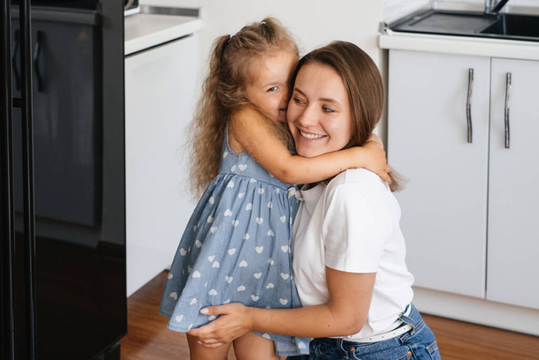 Child whispering something on Mothers ear at home on the kitchen. Kid telling a secret. Toddler girl talking to mom happily asking permission - Zdjęcie, obraz