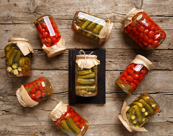 Canned cucumbers and tomato in glass jars on an old wooden background top view. Canned cherry tomatoes and carnishons in jars with kraft lids on a wooden background. - Foto, Bild