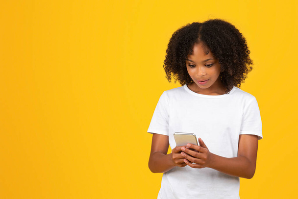 Gelukkig geschokt krullend tiener zwart schoolmeisje in wit t-shirt leest bericht op smartphone, geïsoleerd op gele achtergrond, studio. Studie app, goed nieuws, sociale netwerken, advertentie en aanbod - Foto, afbeelding
