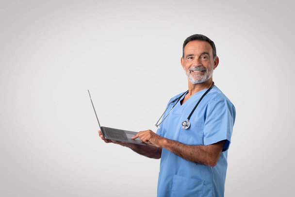 Szczęśliwy biały dojrzały mężczyzna lekarz w niebieskim mundurze piszący na komputerze, odizolowany na szarym tle, studio. Leczenie chorób, konsultacje zdalne, opieka zdrowotna, medycyna i chirurg - Zdjęcie, obraz