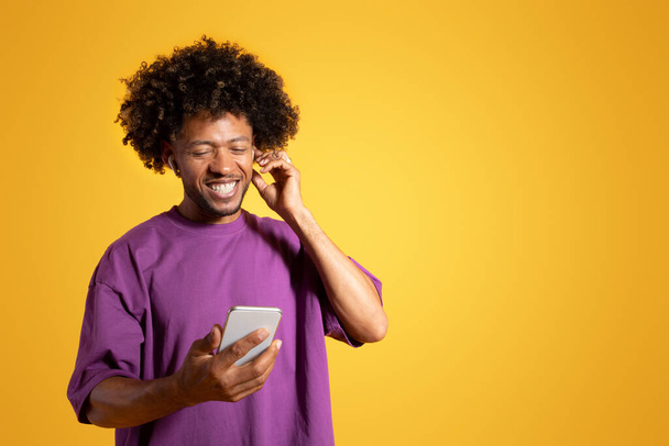 Cheerful black adult curly man in purple t-shirt and wireless headphones looks at phone, isolated on orange background, studio. Listen music for relax, audio app, ad and offer - Foto, immagini
