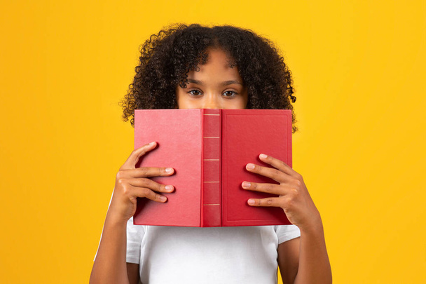 Serious smart curly teenager black schoolgirl in white t-shirt looks from behind book, isolated on yellow background, studio, close up. Education, study, hobbies and homework - Fotografie, Obrázek