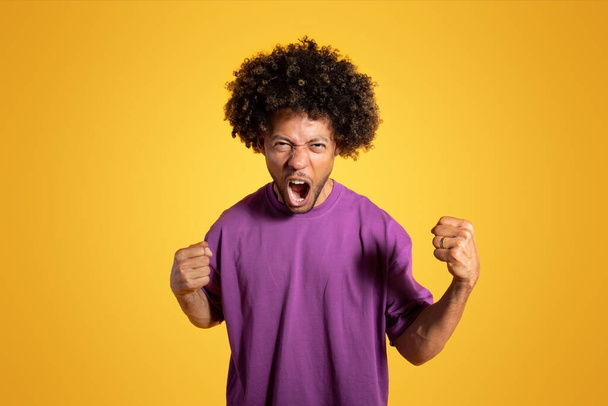 Злий агресивний невдоволений чорний дорослий кучерявий чоловік у фіолетовій футболці з відкритим ротом кричить, ізольований на помаранчевому фоні, студія. Погані новини, емоції, стрес, реклама та пропозиція
 - Фото, зображення