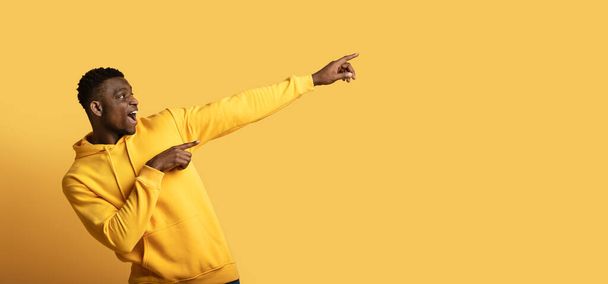 Excited emotional young african american guy in yellow hoodie showing empty space for advertisement, black man pointing aside on yellow studio background, web-banner, panorama - Фото, изображение