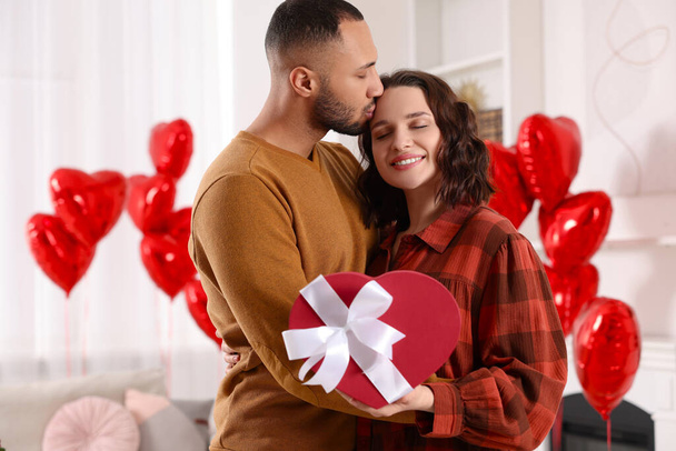 Happy couple celebrating Valentine's day. Beloved woman with gift box in room decorated with heart shaped air balloons - Foto, Imagen
