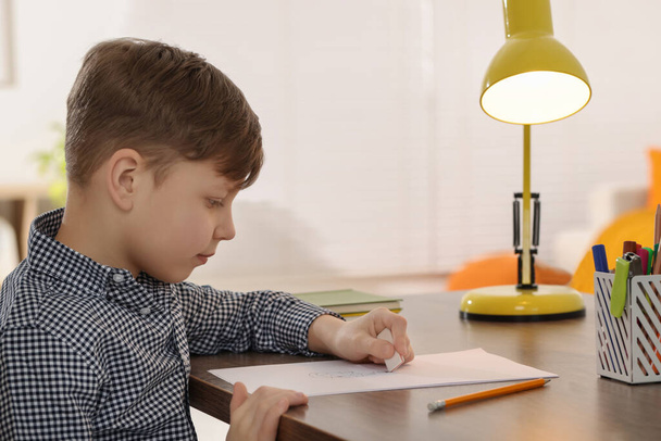 Little boy erasing mistake in his notebook at wooden desk indoors - 写真・画像