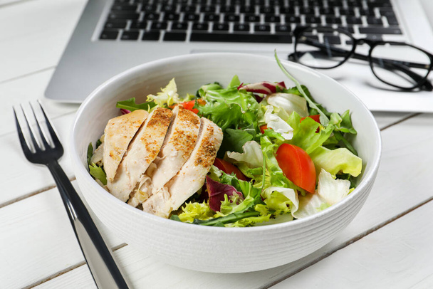 Bowl with tasty food, fork, laptop and glasses on white wooden table, closeup. Business lunch - Фото, изображение