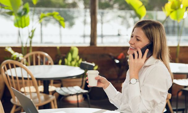 Side view of young businesswoman in formal clothes sitting at table with cup of coffee and talking on smartphone at cafe during daytime - Valokuva, kuva