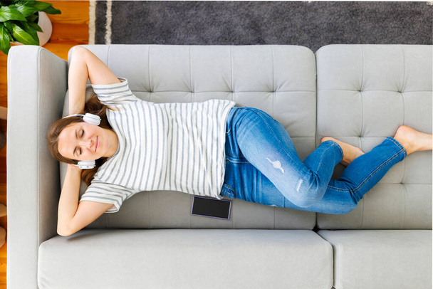 Friedliche Frau in modernen drahtlosen Kopfhörern, die sich auf einer bequemen Couch entspannen und Musik hören. Glückliche ruhige Frau mit Kopfhörern entspannt auf einem bequemen Sofa, genießen Sie hochwertigen Sound - Foto, Bild