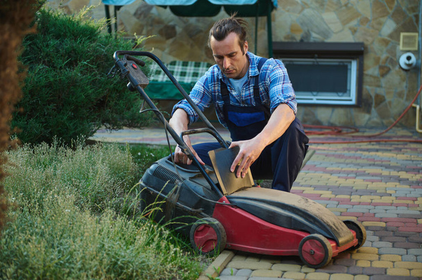 Yakışıklı İspanyol işçi, mavi bahçıvan üniformalı bahçıvan, filtredeki kesilmiş çimenleri temizliyor, daha fazla gübrelemek için, bahçedeki çim biçme makinesini şarj ediyor. - Fotoğraf, Görsel