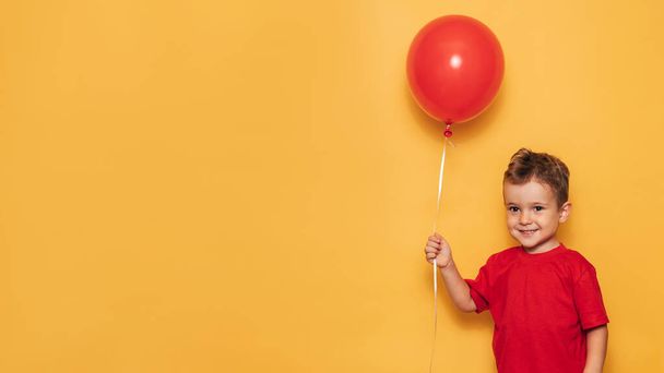 Un niño caucásico feliz aislado sobre un fondo amarillo brillante sostiene un globo rojo en sus manos. Un lugar para su texto o anuncio - Foto, imagen