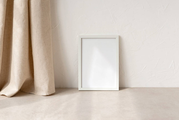 Empty white picture frame mockup on a neutral beige table and wall background, linen curtains and soft sunlight shadow pattern. Minimalist modern home office interior workspace, art, poster display - Photo, Image