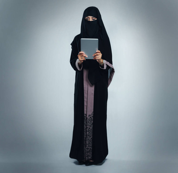 Utrzymując jej styl życia cyfrowy. Studio zdjęcia młodej kobiety noszącej burkę i korzystającej z tabletu cyfrowego na szarym tle - Zdjęcie, obraz