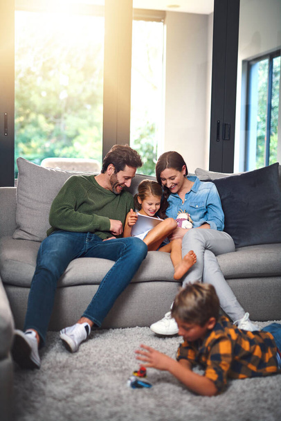 Bonding en browsen. een moeder, vader en dochter die samen op de bank een tablet gebruiken terwijl hun zoon in de woonkamer thuis op de vloer speelt - Foto, afbeelding