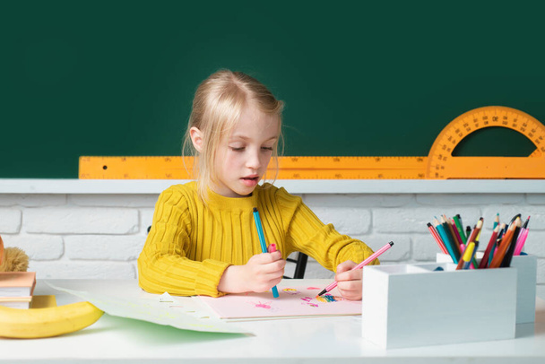 Back to school. Cute pupil girl drawing at the desk. Genius child, knowledge day. Kids education and knowledge. Child in the class room with blackboard on background. Kid girl from primary school - Fotoğraf, Görsel