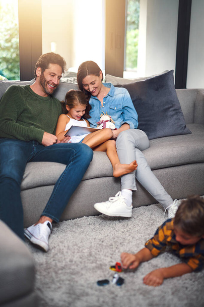 He loves his toys and she loves her cartoons. a mother, father and their daughter using a tablet together on the sofa while their son plays on the floor in the living room at home - Photo, image