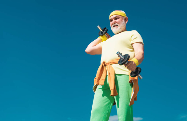 Senior sportman exercising with lifting dumbbell on blue sky background. Isolated, copy space. Portrait of senior man holding dumbbell. Healthcare cheerful lifestyle - Foto, Imagem