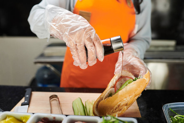Руки в перчатках молодой работницы уличного фургона быстрого питания наливают соус в аппетитный хот-дог с различными ингредиентами внутри - Фото, изображение