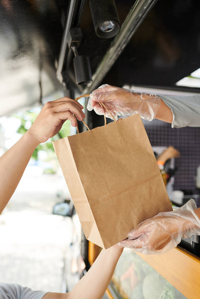Nahaufnahme der Hände junger Konsumenten, die Papiertüten mit Fast Food von einer Arbeiterin des Streetvan-Kochens und des Verkaufs von Hotdogs entgegennehmen - Foto, Bild