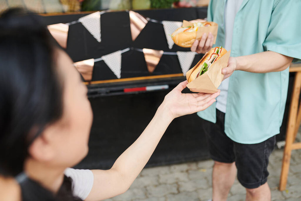 zbliżenie młody kobieta biorąc świeże apetyczny hot dog zawinięte w papier z ręki jej chłopak w casualwear stoi z przodu - Zdjęcie, obraz