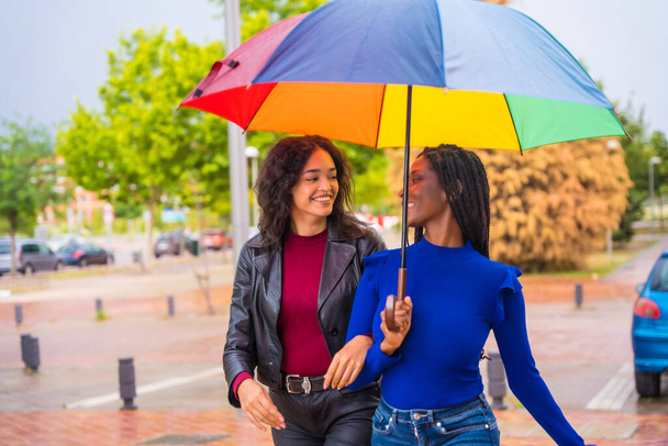 Retrato de mujeres amigas multiétnicas riendo con un paraguas bajo la lluvia en una ciudad - Foto, imagen
