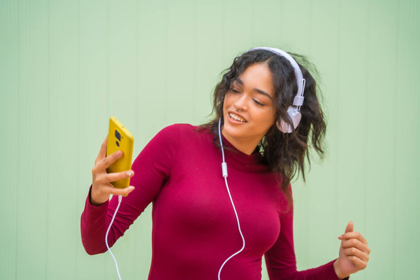 Portret van een latijnse vrouw met een glimlachende koptelefoon, technologieconcept op een groene achtergrond, luisteren naar muziek - Foto, afbeelding