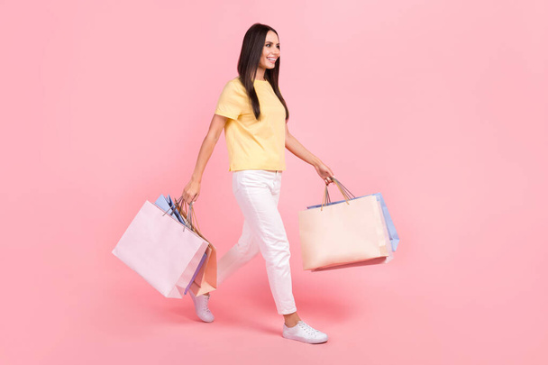 Full length photo of pretty cute lady wear yellow t-shirt walking holding bargains looking empty space isolated pink color background. - Fotoğraf, Görsel
