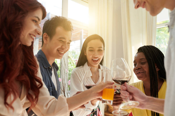 Joyeux jeunes amis interculturels claquant avec leurs boissons à la fête à la maison devant la caméra tout en profitant de la célébration et en s'amusant - Photo, image