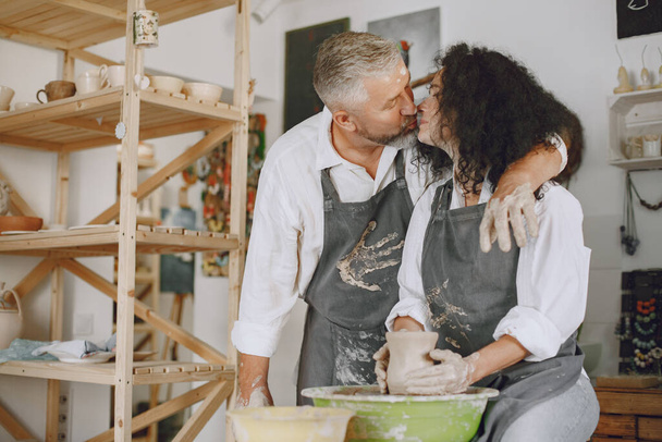 Mutual creative work. Adult elegant couple in casual clothes and aprons. People creating a bowl on a pottery wheel in a clay studio. - Photo, image