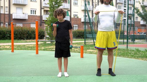 Italy, Milan - Father and son kid child 9 years old do gymnastics gym in a park with weight equipment area - physical exercises sport activities and summer preparation diet - Photo, Image