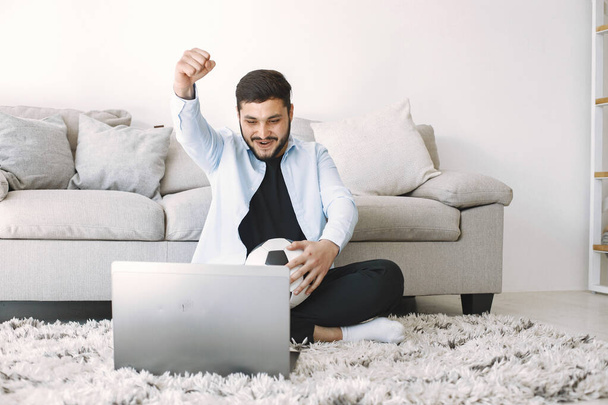 Young excited bearded man on a floor near the couch while watching football match on laptop. Man holding his hand in the air. Man in blue shirt sitting on carpet. - Foto, Imagem