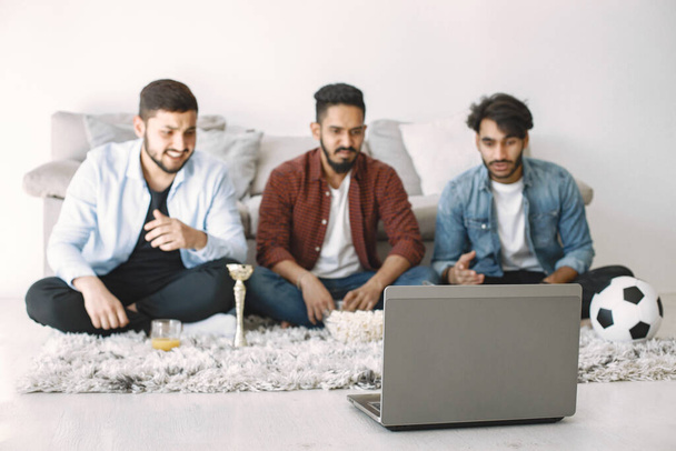 Three young excited male friends sitting on a floor while watching football match on TV. Focus on a grey laptop. Men wearing shirts and t-shirts. - Fotó, kép
