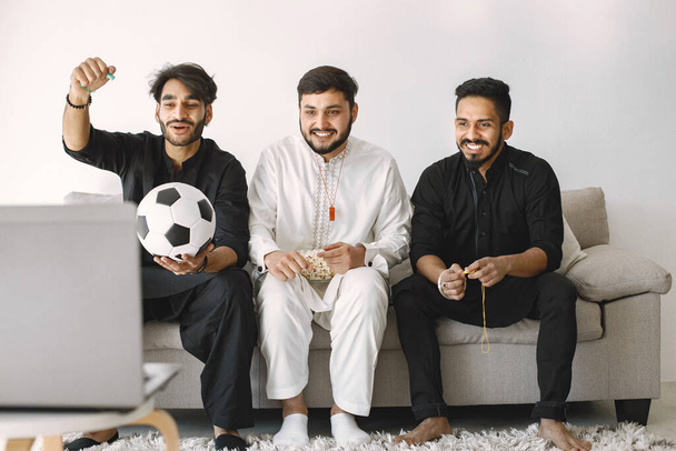 Three indian male friends sitting on a coach while watching football match on laptop. Men have a ball and eating popcorn. Men wearing black and white indian clothes. - Foto, afbeelding