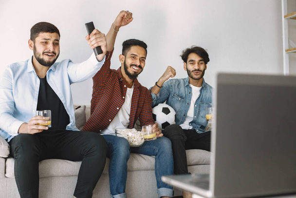 Troje młodych, podekscytowanych przyjaciół siedzących w autokarze i oglądających mecz piłki nożnej na laptopie. Mężczyźni piją sok i jedzą popcorn. Mężczyźni w koszulkach i koszulkach. - Zdjęcie, obraz