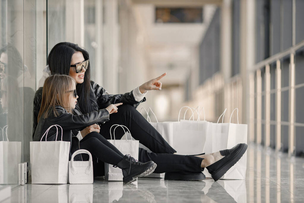 Mother and daughter on shopping. Young pretty clients standing in front of glasses wall holding shopping paper bags. Brunette woman with her daughter wearing black clothes. - Valokuva, kuva