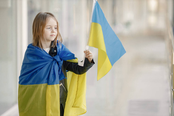 Little girl with ukrainian flag posing for a photo. Young pretty girl standing in front of glass wall. Girl supporting Ukraine. - Foto, imagen