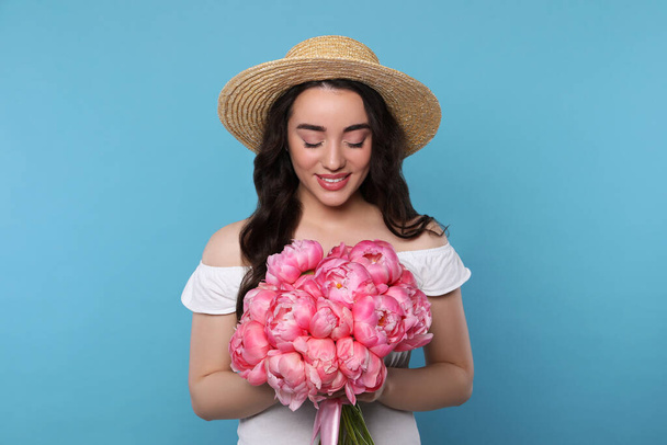 Beautiful young woman in straw hat with bouquet of pink peonies against light blue background - Фото, изображение