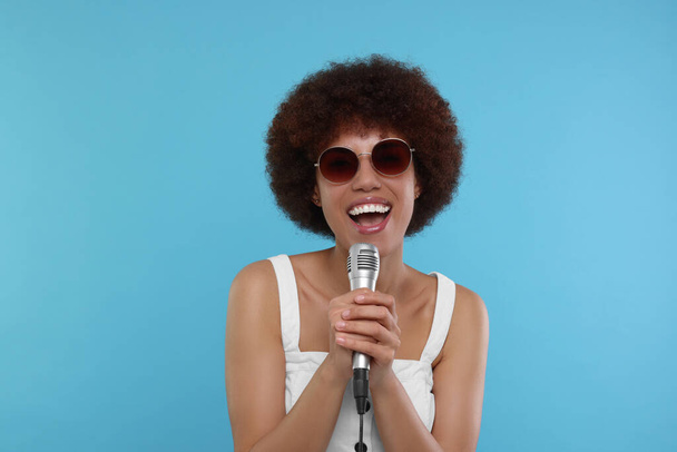 Curly young woman in sunglasses with microphone singing on light blue background - Фото, изображение