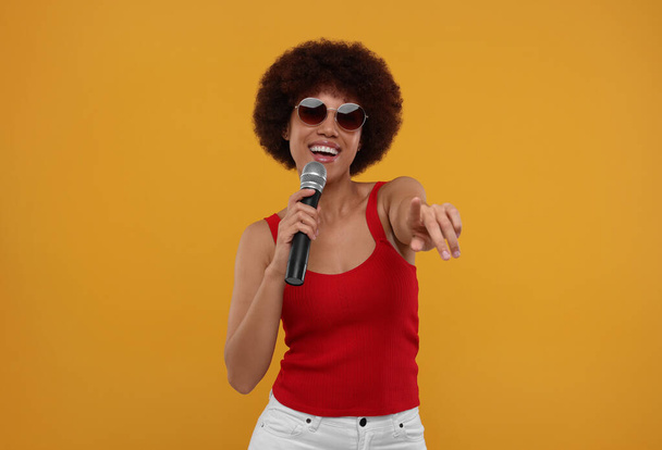 Curly young woman in sunglasses with microphone singing on yellow background - 写真・画像