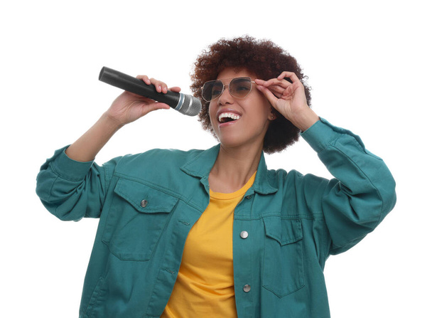 Кудрявая молодая женщина в солнечных очках с микрофоном поет на белом фоне - Фото, изображение